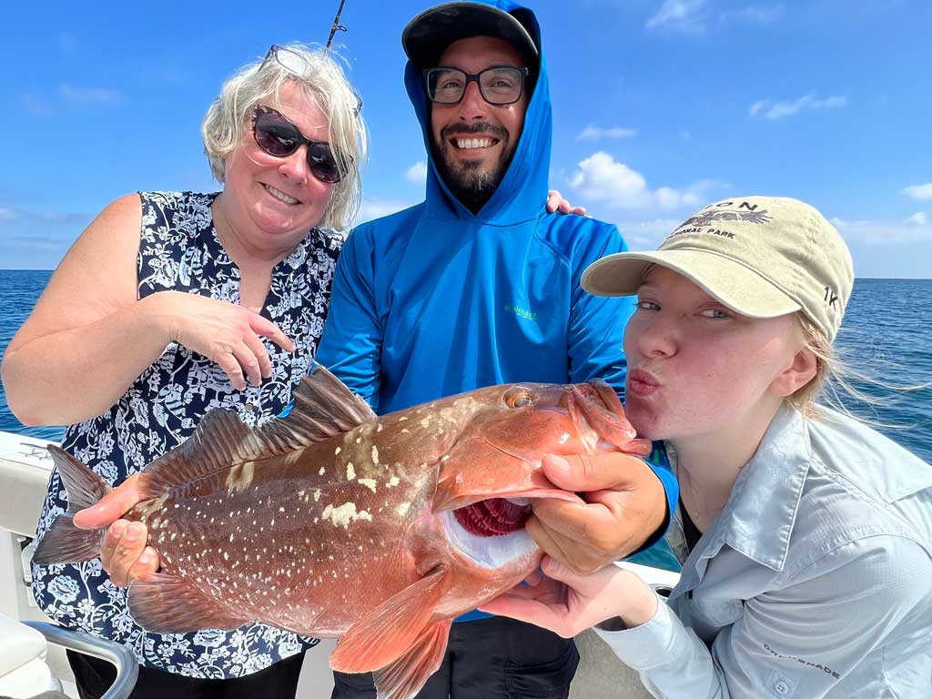 A photo of two females and one male angler posing on a charter boat with a Grouper caught while bottom fishing nearshore