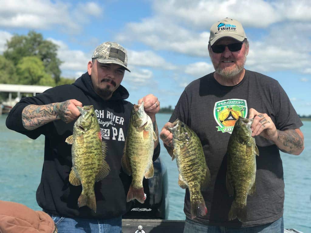 Two anglers standing side by side on a boat wearing hats and with visible tattoos, as they hold two Smallmouth Bass each that they caught fishing on Lake St. Clair.