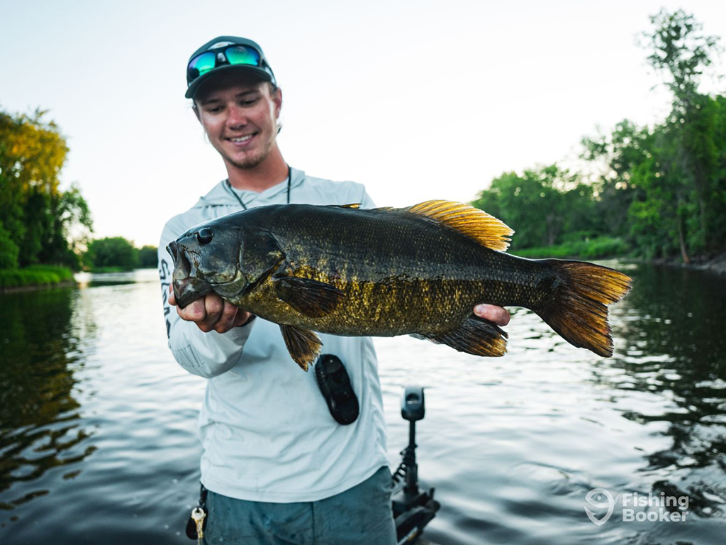 A youthful angler in a baseball cap holds a large Smallmouth Bass aboard a fishing charter towards the end of the day on a calm body of water in Minnesota