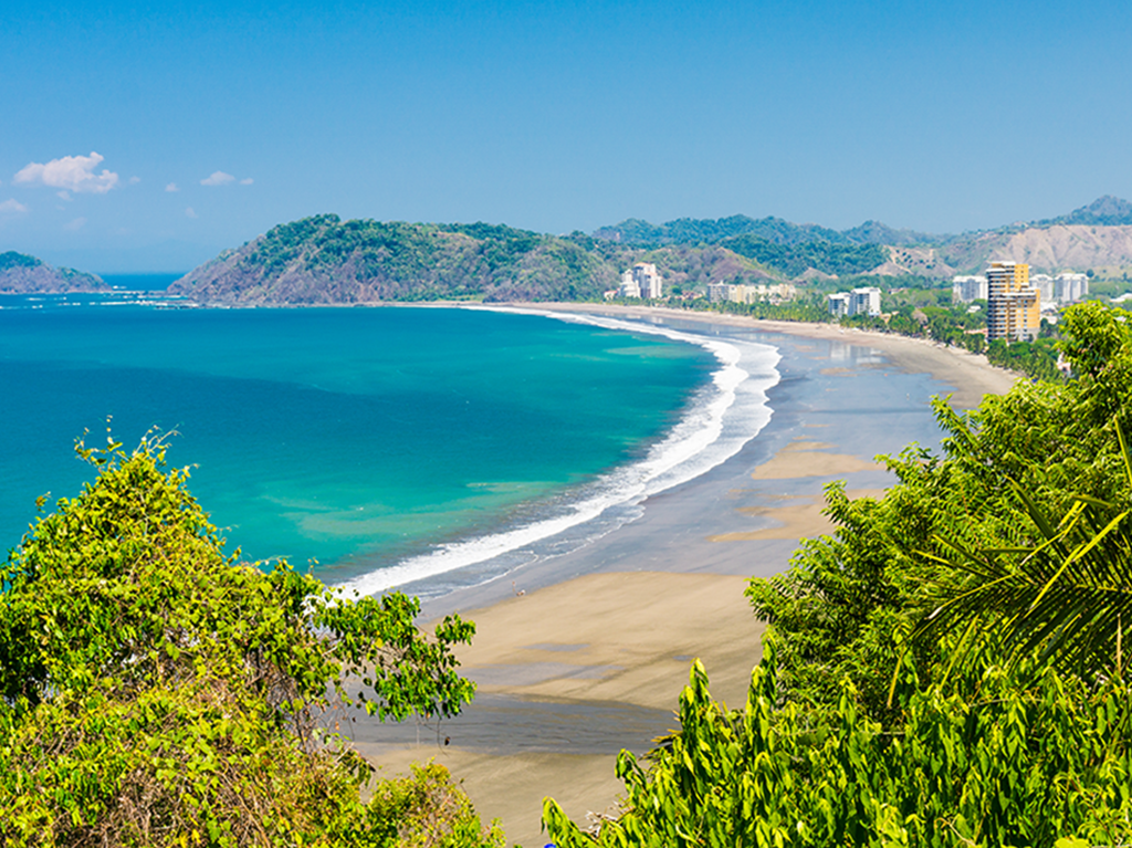 A view from a hill along the beach in Jaco, Costa Rica, on a clear day, with the beach on the right of the image and a hill visible in the distance