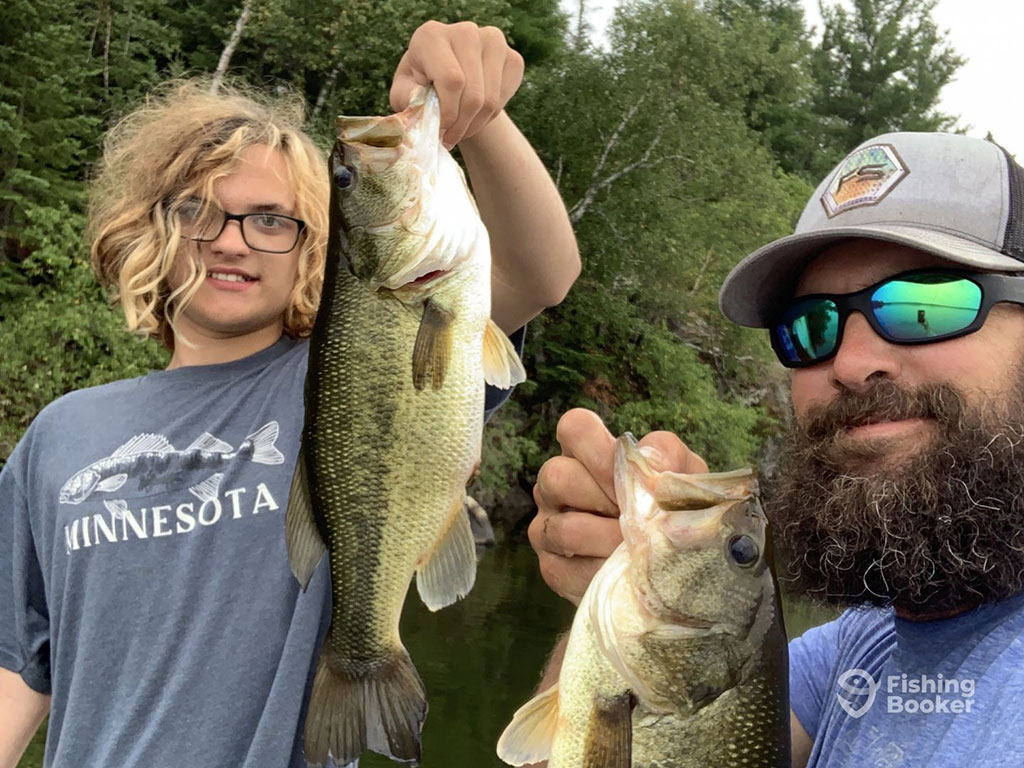 A young boy with curly blonde hair and a man with a beard hold up a Largemouth Bass each while taking a selfie aboard a fishing charter in Minnesota