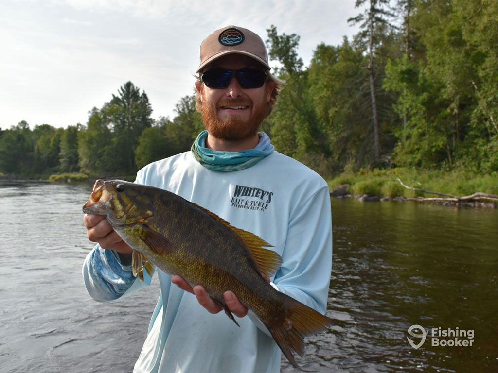 A man in a baseball cap and sunglasses standing on a fishing charter in Minnesota holding a Smallmouth Bass on a clear day, with a green shoreline visible behind the water behind him