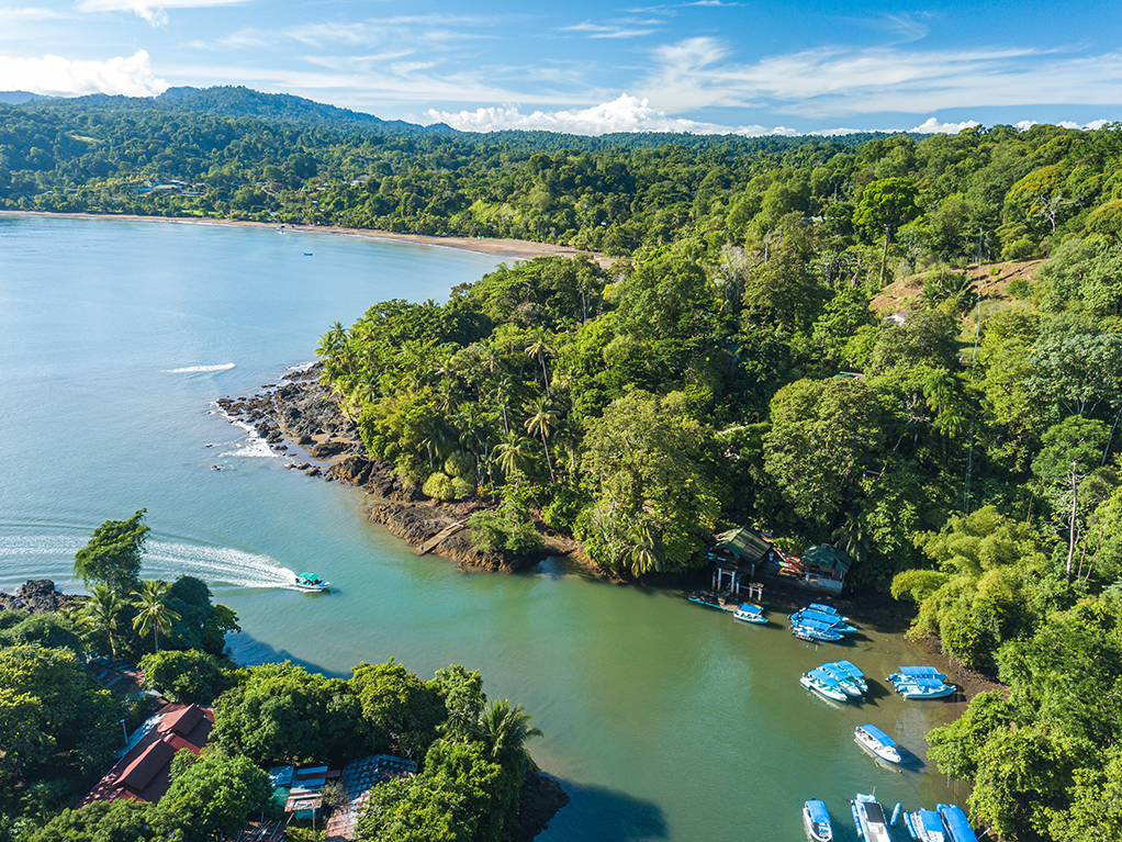 An aerial view down to a small bay on the Osa Peninsula, Costa Rica, on a sunny day, with blue boats visible in the water next to a lush, green shoreline