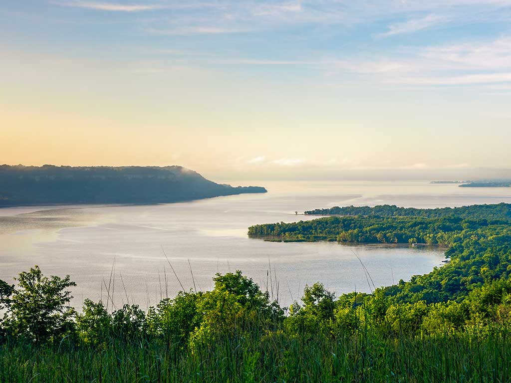 A view from a hill of the Mississippi River as it turns into Lake Pepin in Minnesota at dawn on a sunny day, with the sun just about to creep up over a hill in the distance