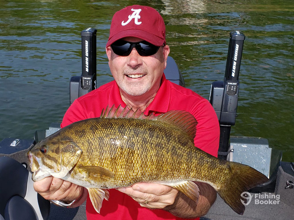 A man in a red shirt and hat holding up a Smallmouth Bass to the camera on a sunny day with the water visible behind him