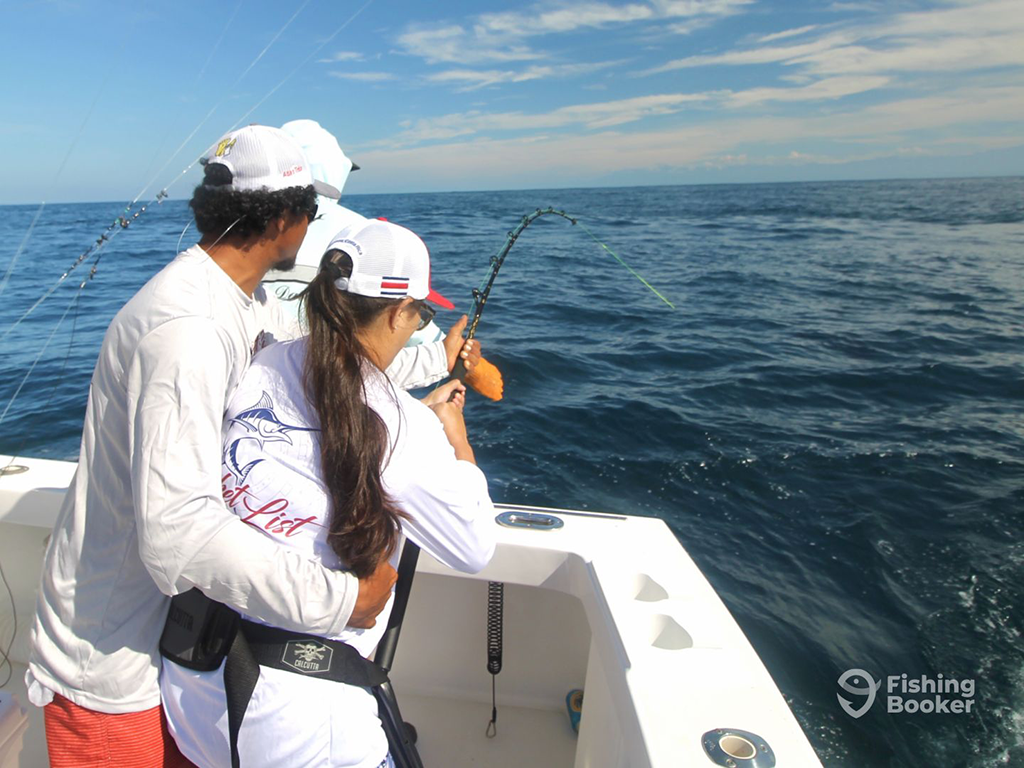 A view from behind of a man with his arm around the waist of a woman, as he helps her reel in a big fish aboard a fishing boat in Costa Rica