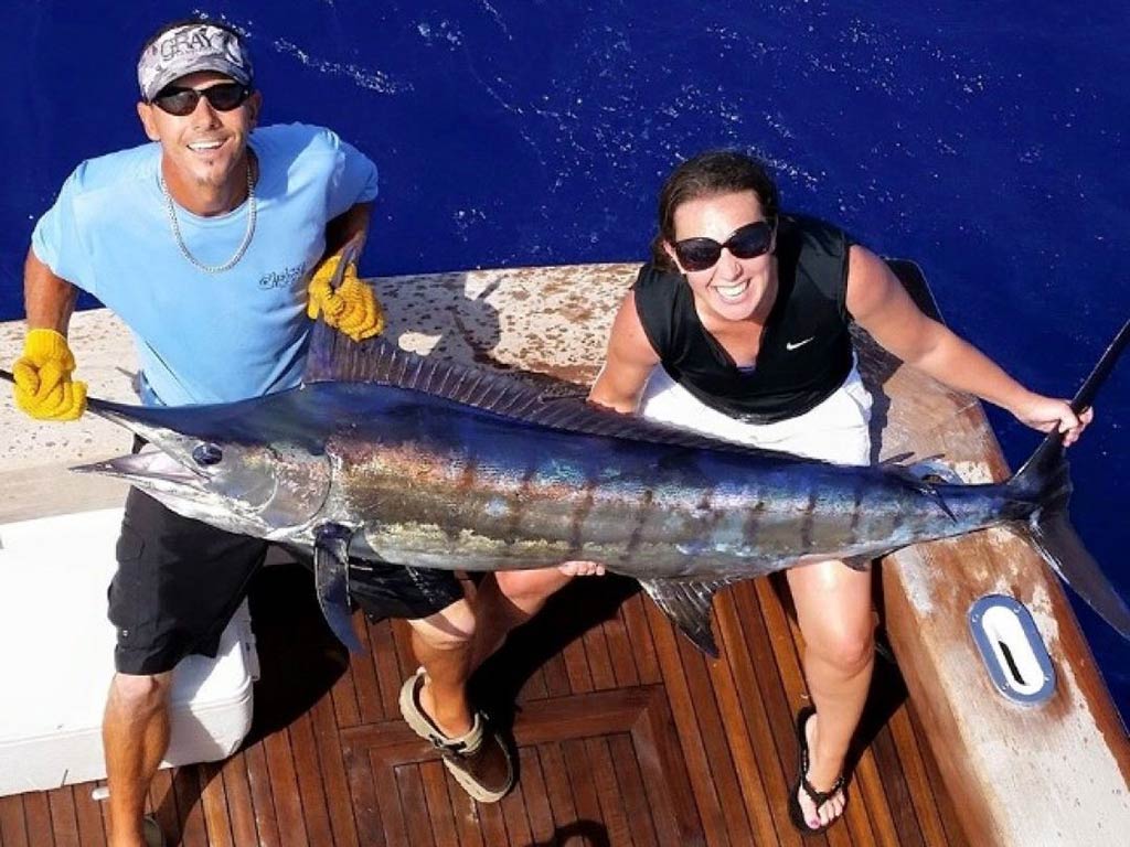 A view from a flybridge down to the deck of a sportfishing boat in Key West, with two anglers – one male, one female – smiling as they hold a large Marlin with the water behind them