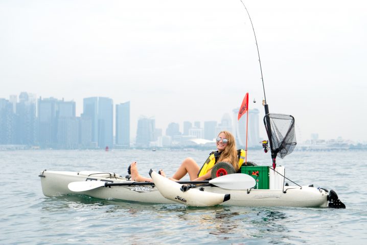 A blond woman in sunglasses sitting in a white fishing kayak on the sea with a cityscape behind her in the distance