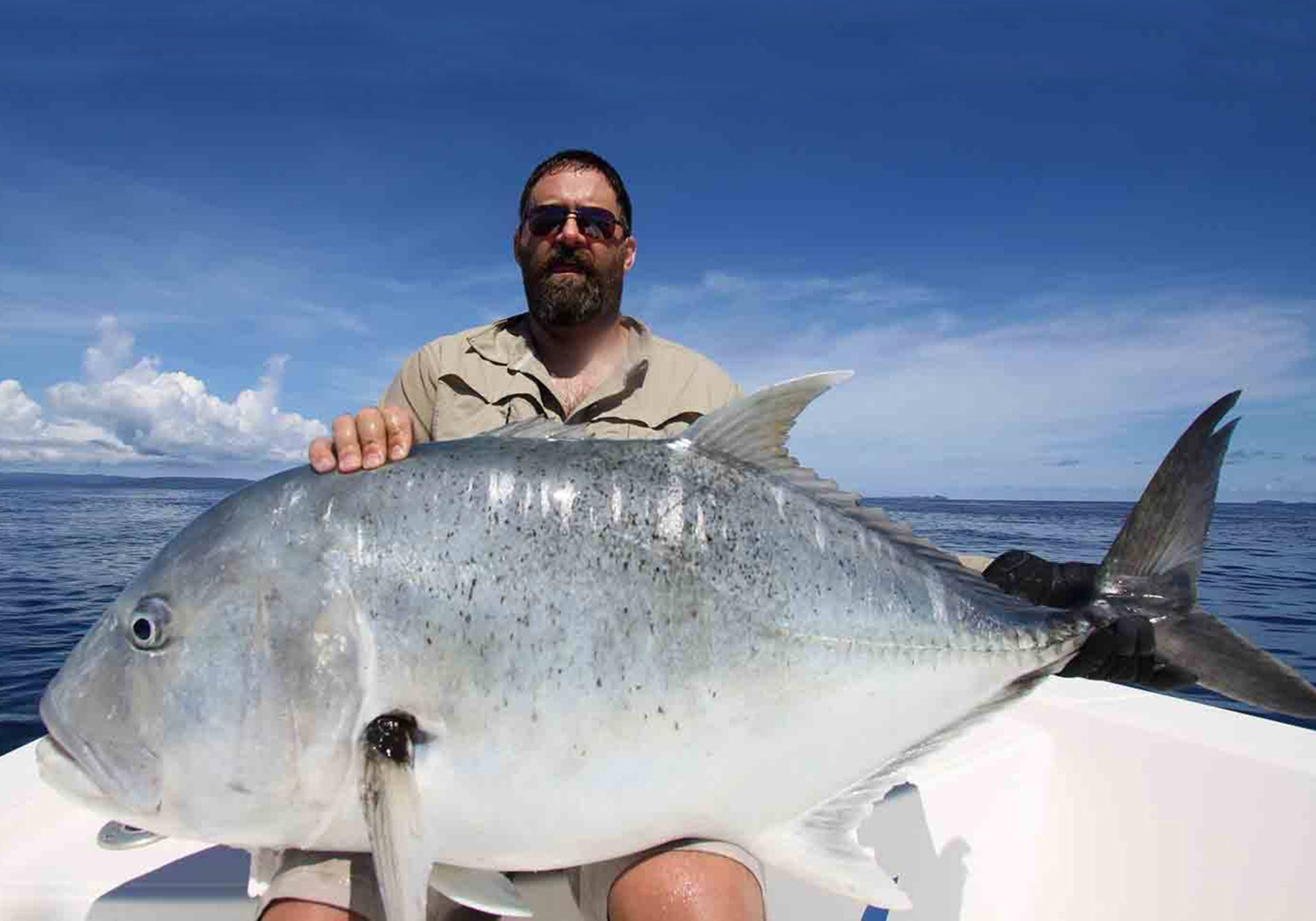 A male angler with a large Giant Trevally fish on his lap.