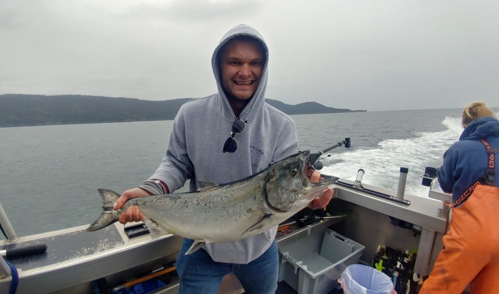 a smiling angler holding a large Salmon on a fishing boat in Alaska, showing that fishing is a great guilt-free activity to enjoy during the covid pandemic