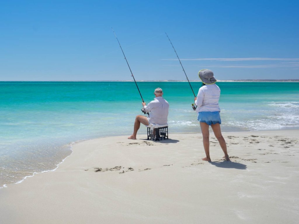 Two people standing on a white-sand beach and fishing