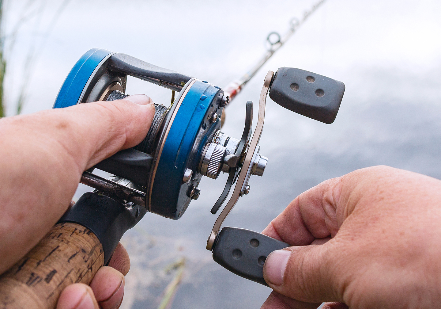 A baitcaster fishing reel being held near some water.