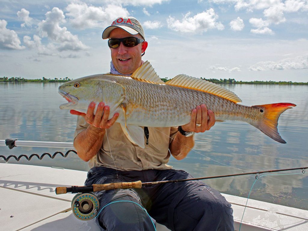A fly angler with his rod in his lap holding a big Redfish while sitting on a boat