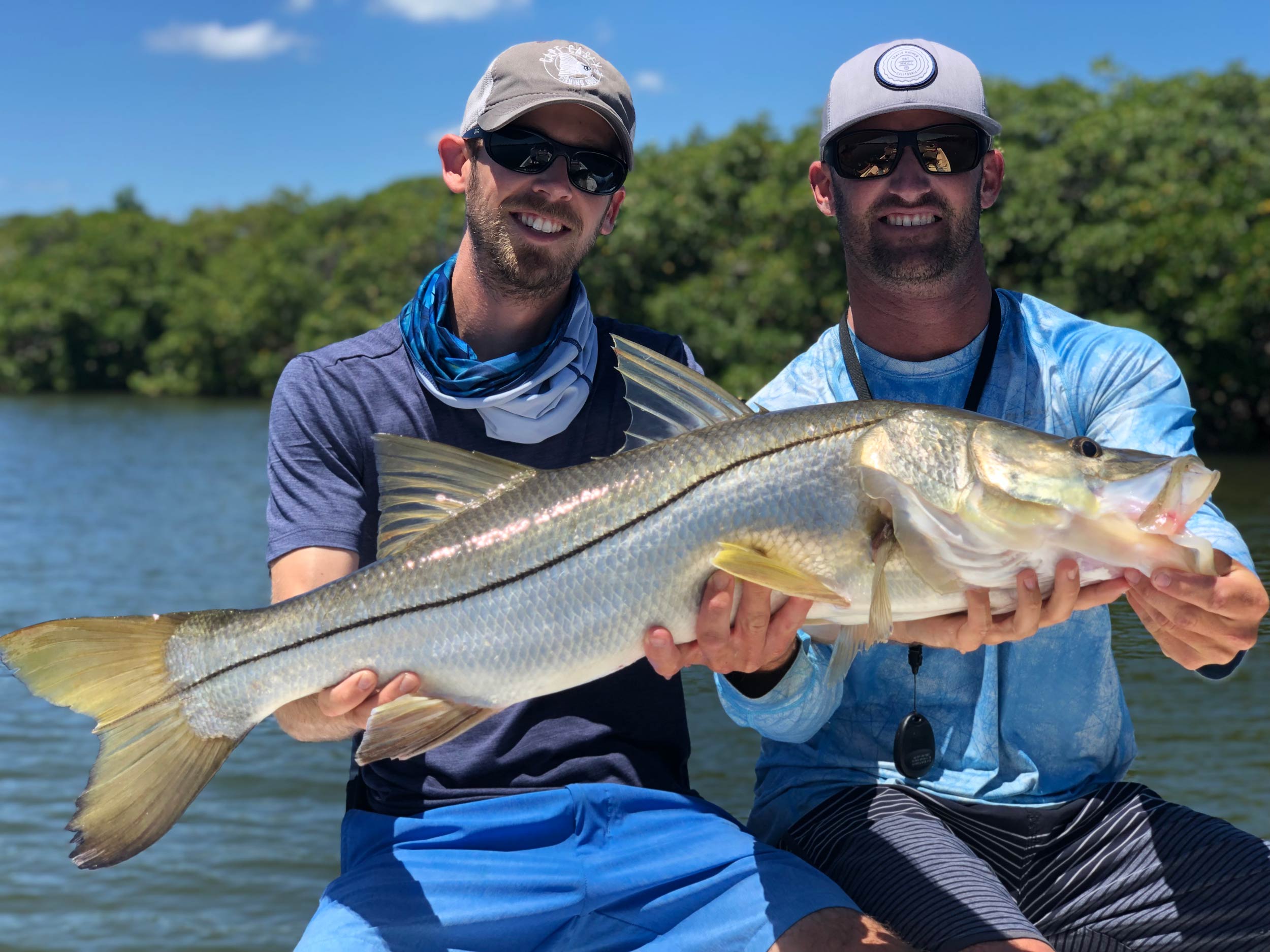 Two smiling anglers sitting on a boat with a large Snook in their hands with trees in the background