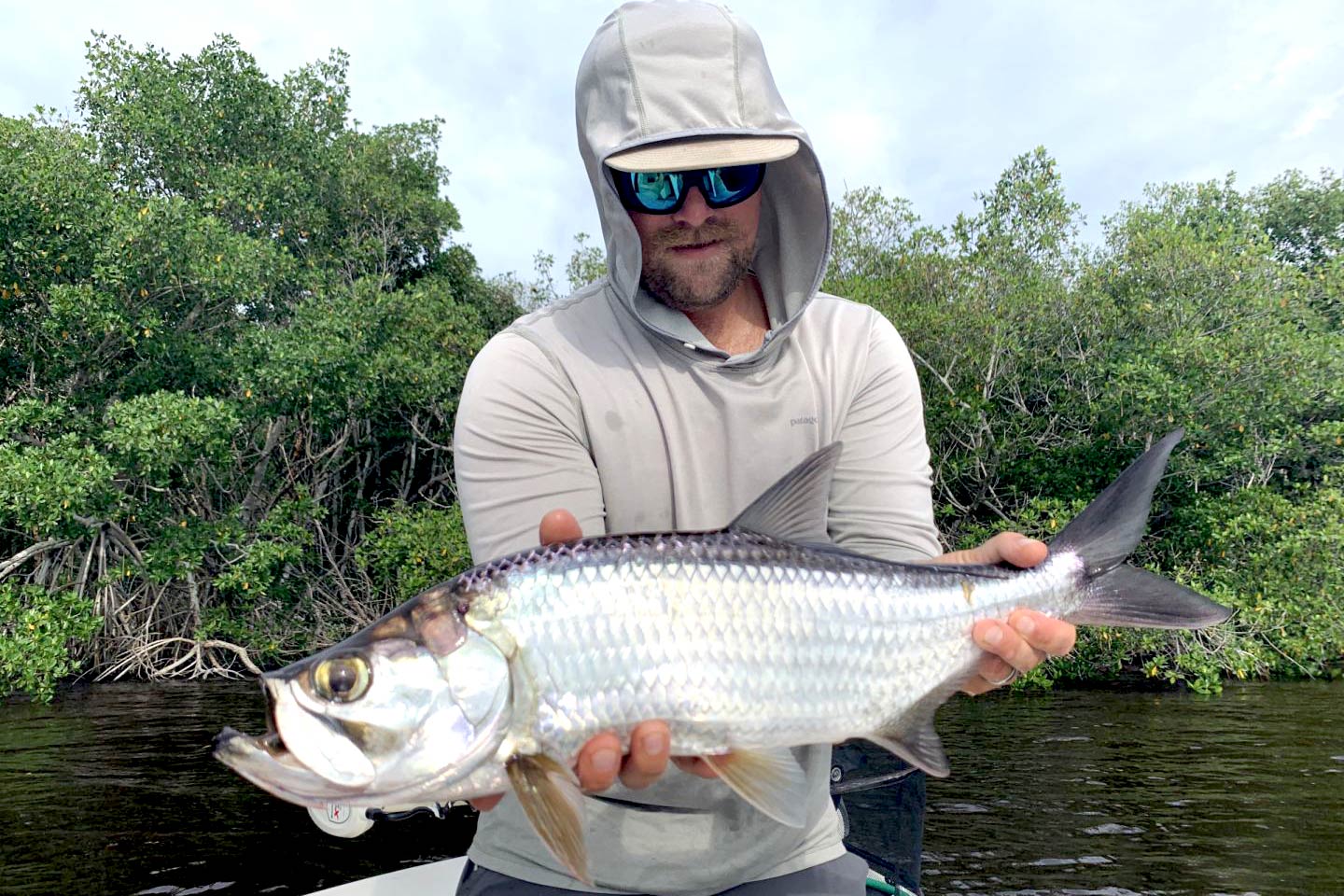A hooded angler holing a Tarpon on a boat with trees in the background
