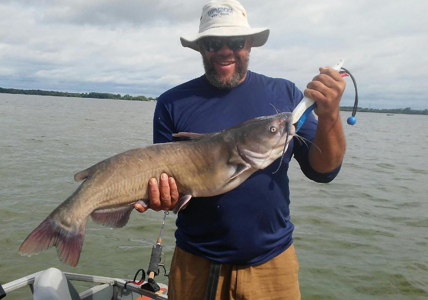 A smiling angler in a hat holding a Channel Catfish with water in the background