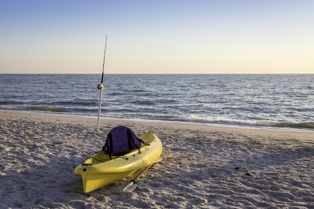 A yellow fishing kayak on a beach, with a surf fishing rod near the water behind it