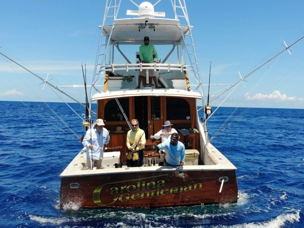 A view of the back of an offshore sportfishing boat, with four anglers fishing and the captain looking down from the flybridge on a sunny day  