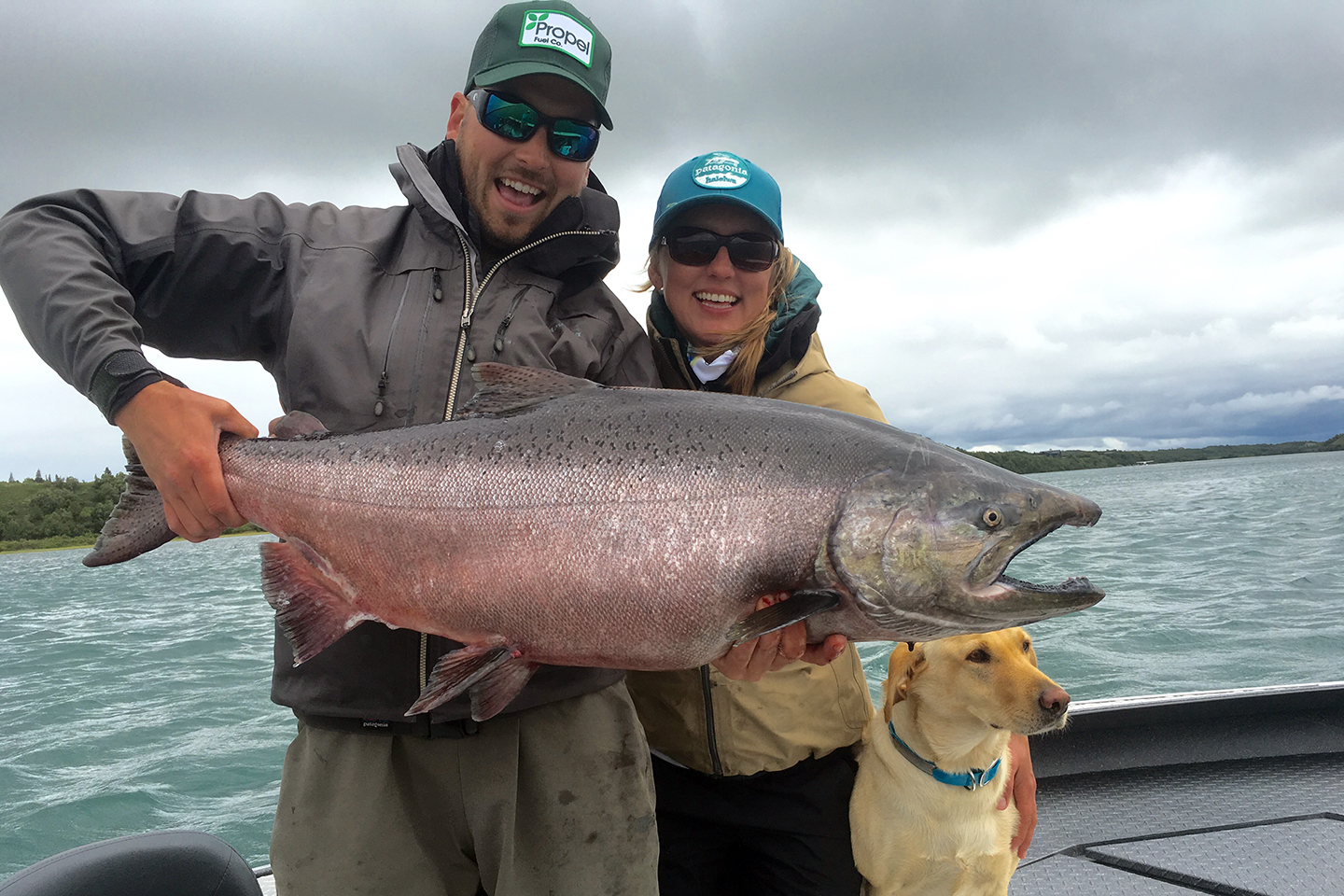 A photo of a couple posing with their dog and a large Chinook Salmon on a boat during a cold fishing season