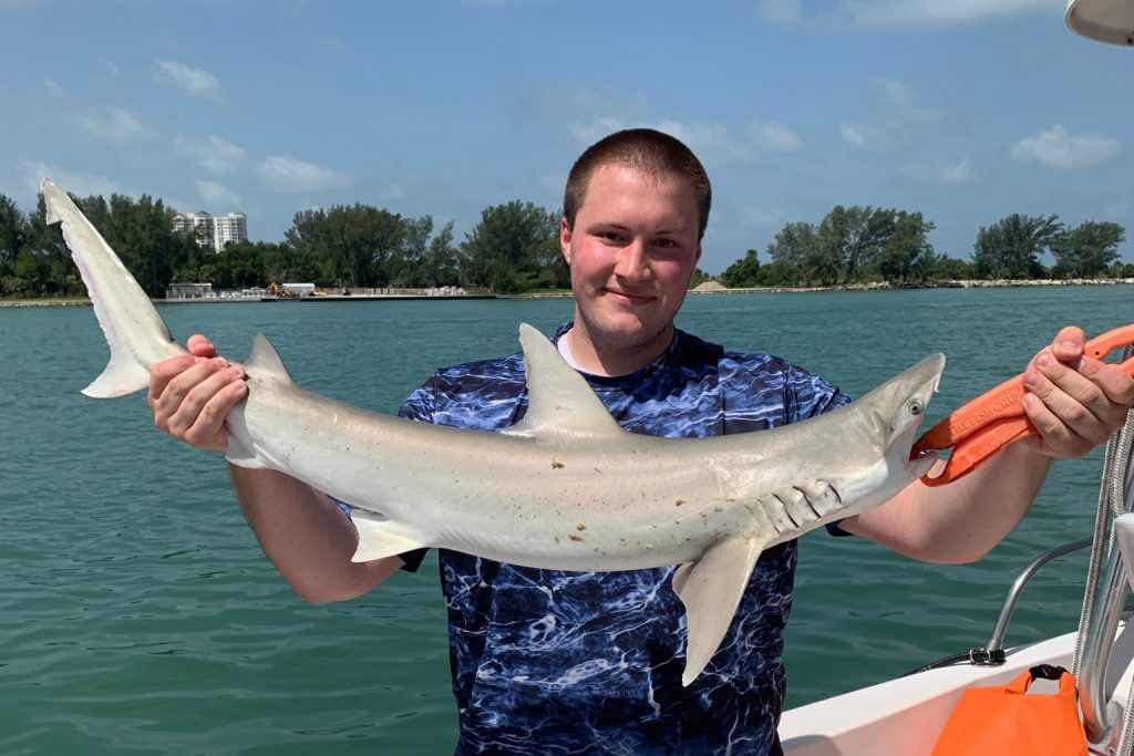 A man holding a Shark on a boat, with the shoreline in the distance behind him