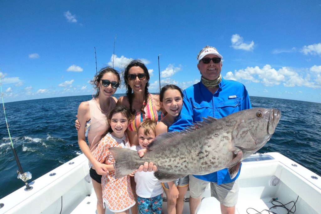 A family stand aboard a charter vessel in Fort Morgan holding a large Grouper with the Gulf behind them
