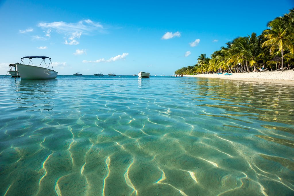 Clear, shallow sea and small boats on the water in Belize, with a yellow sandy beach and tropical trees on the right.