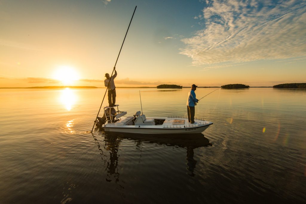 Un bateau de pêche plat sur une eau calme et peu profonde au coucher du soleil. Le capitaine est debout à l'arrière, poussant le bateau avec une perche. Un pêcheur se tient à l'avant avec une canne à pêche.