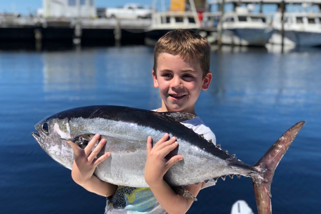 A smiling kid holding a Tuna he caught on a fishing trip