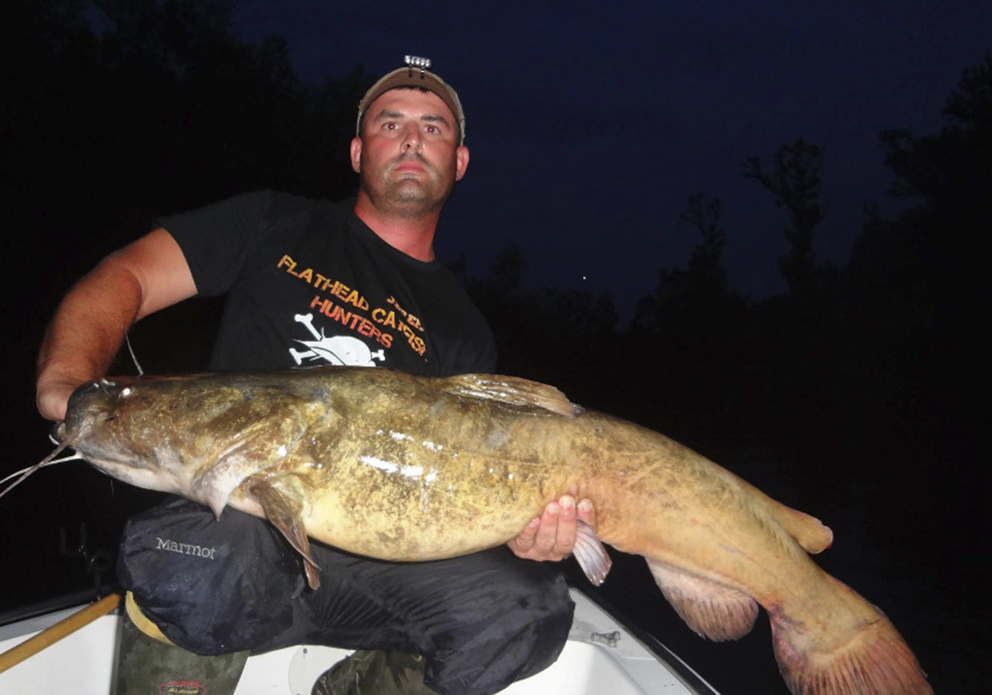 A man on a boat at night holding a big Flathead Catfish