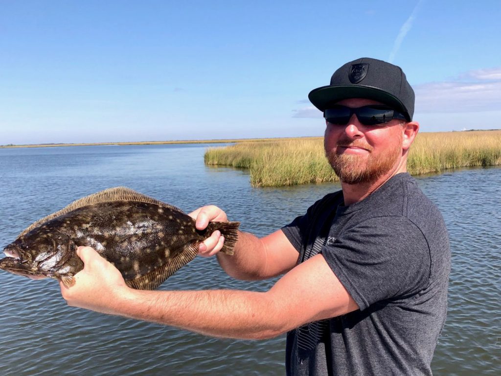 A fisherman holding a Flounder with blue skies and lake Pontchartrain in the background