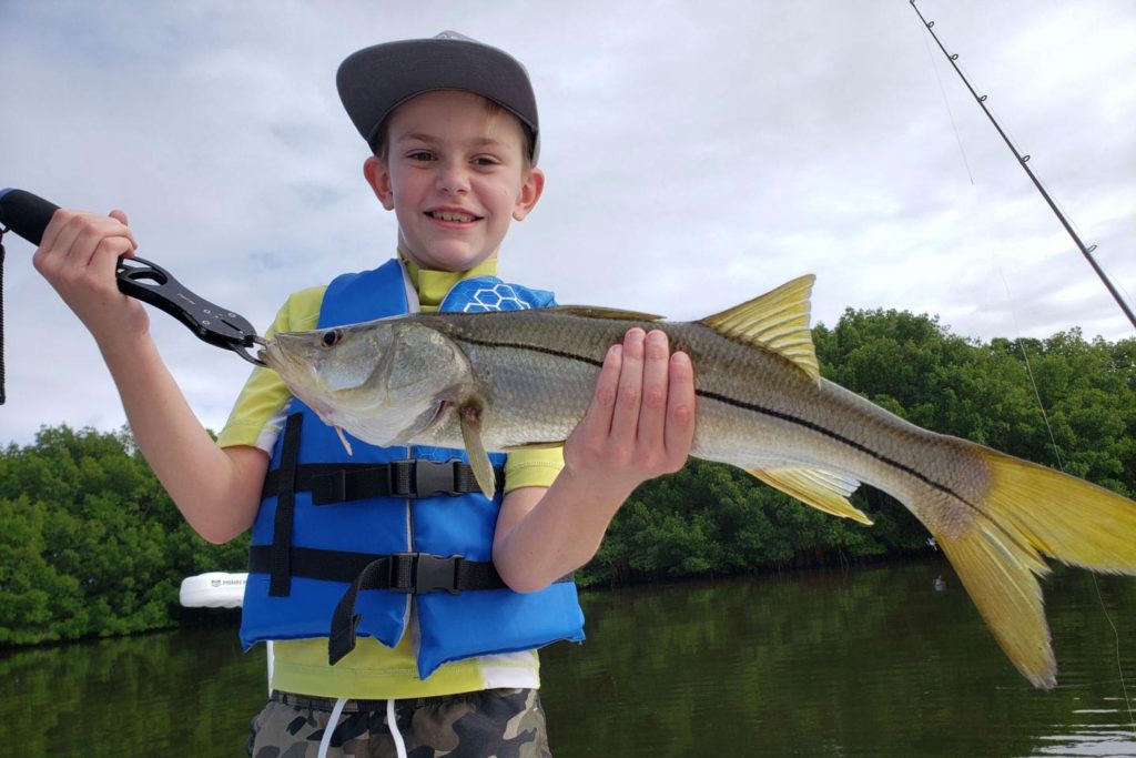 A child holds a Snook on a boat with the marshy waters of a Gulf bay behind him