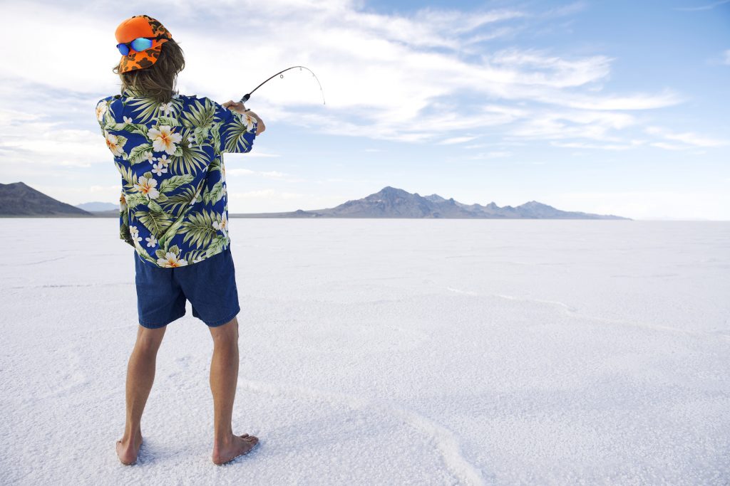 A man in shorts and a Hawaiian holding a rod on a sheet of ice, showing the classic setup for many funny fishing jokes.