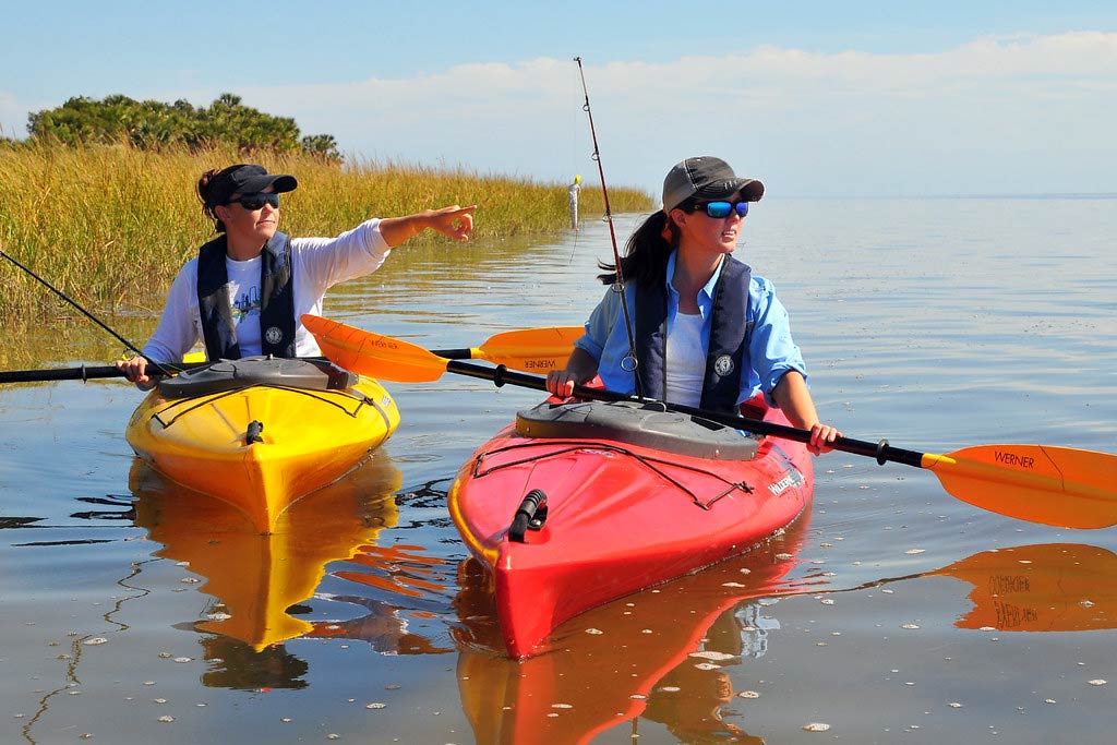 Two women kayaking through the flats with fishing rods, while one points to their left