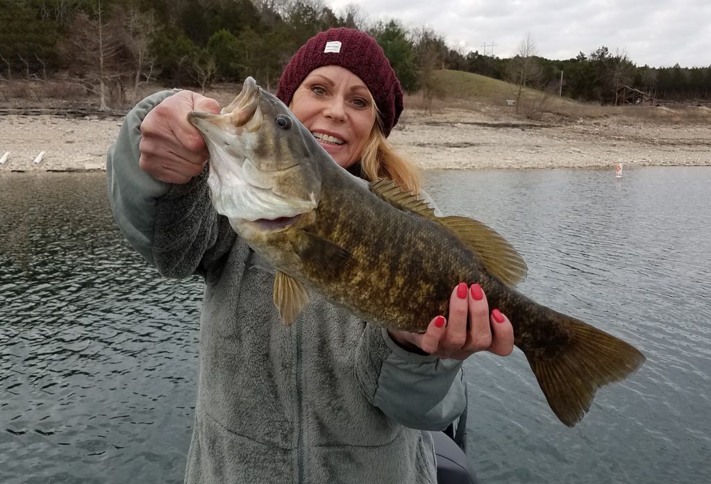 A lady holding a Bass in front of a scenery of water and brush.