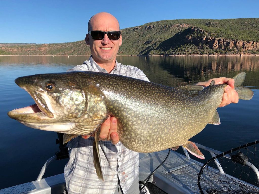 A smiling man in sunglasses holding a big Lake Trout, with Lake Huron in the background