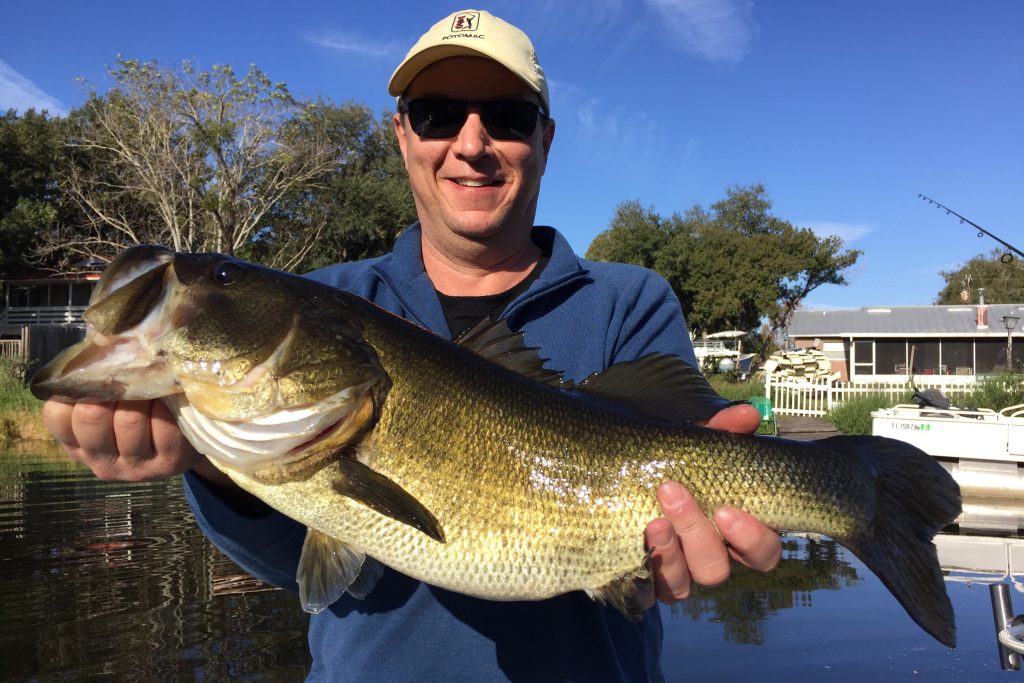 A happy man in a cap, smiling while holding a Largemouth Bass he caught while fishing, with a narrow waterway and a house behind him