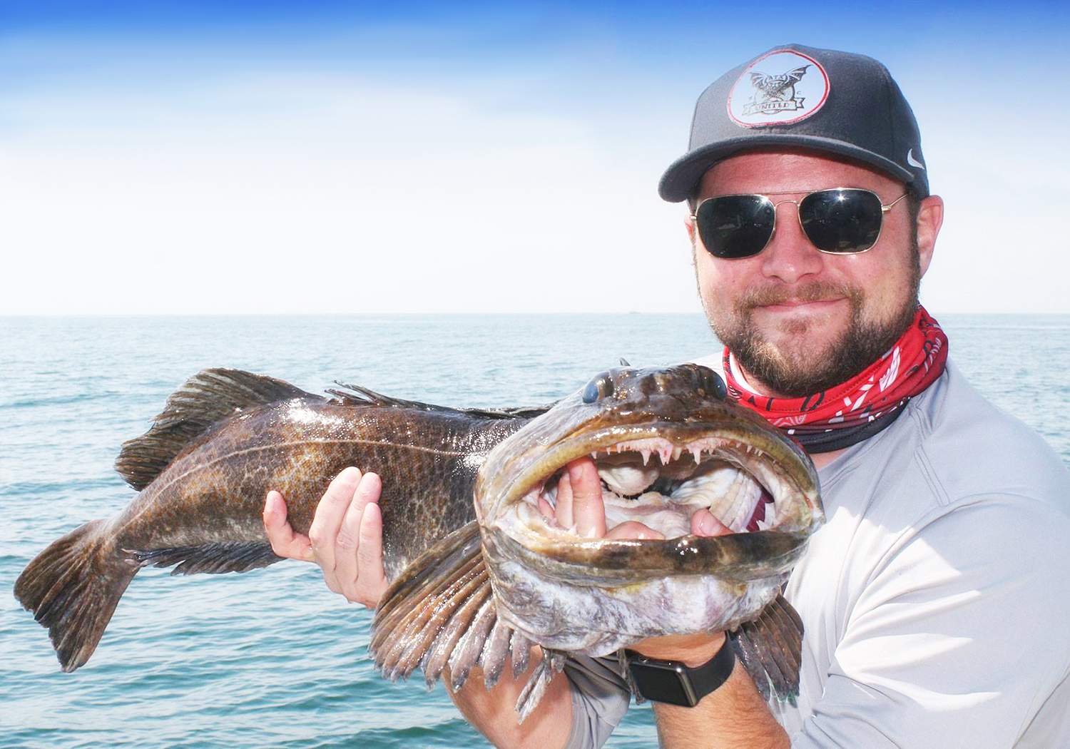 Ein lächelnder Angler, der einen Lingcod hält, mit blauem Himmel und Wasser im Hintergrund