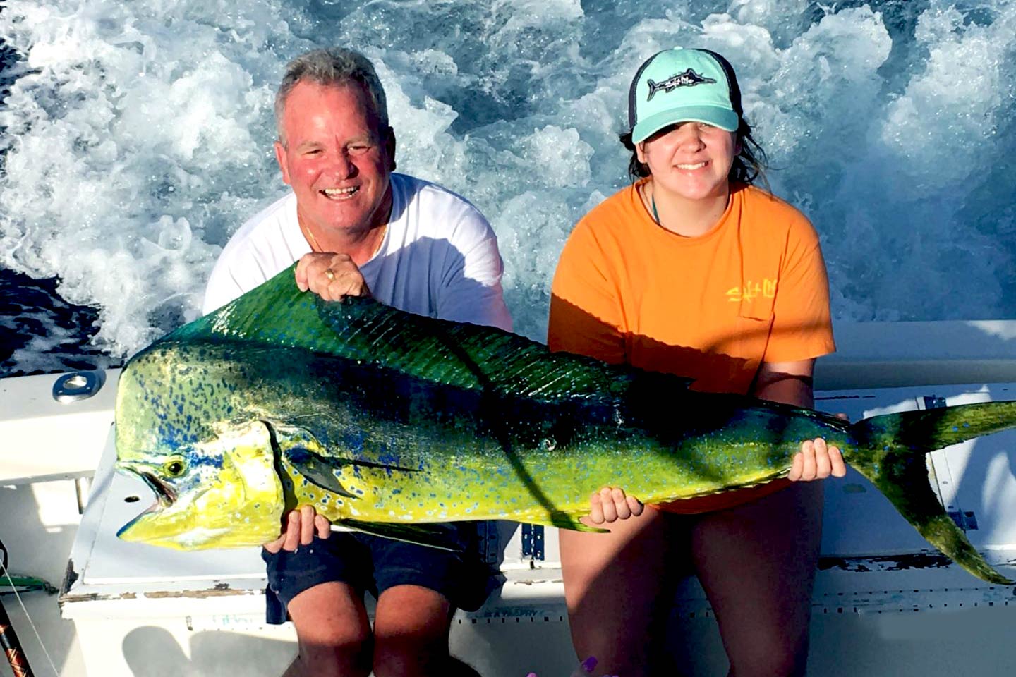 A man and a woman sitting on the back of a boat in Florida and holding a big Mahi Mahi, with the engine creating a wake behind them