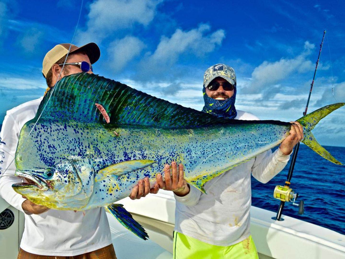 Two men on an offshore sportfishing vessel wearing hats and sunglasses, smiling and holding a large Mahi Mahi