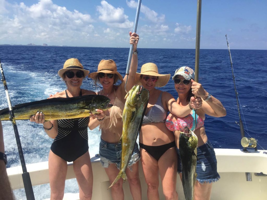 Four women standing on a boat, holding three Mahi Mahi fish with blue water in the background