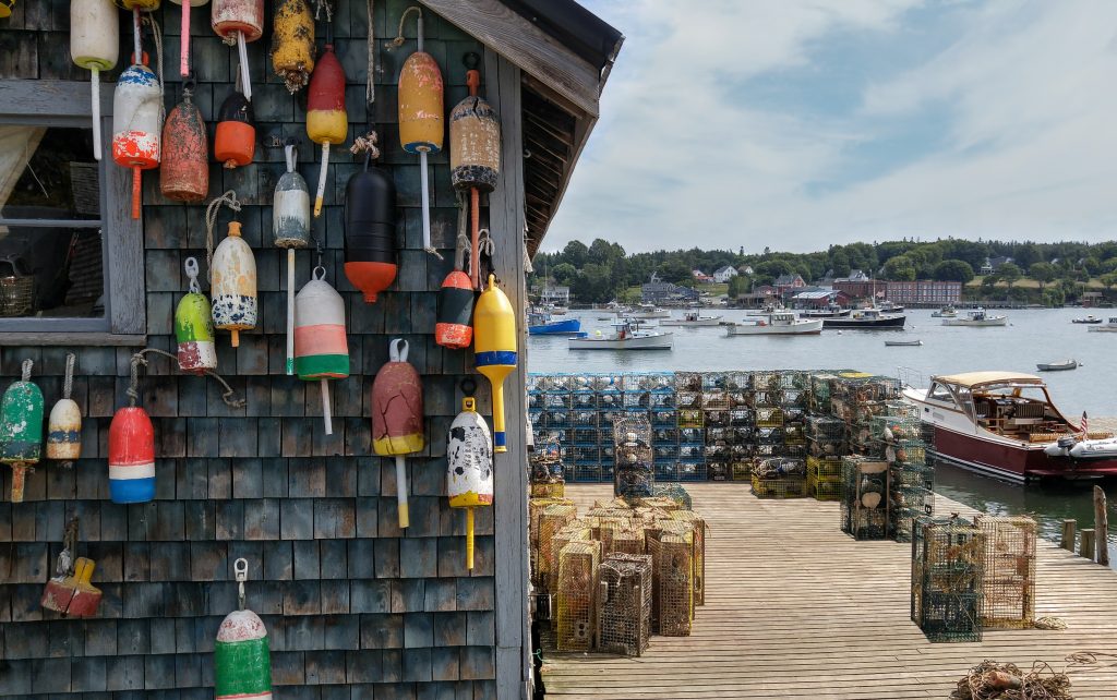 A traditional dock in a fishing village in Maine, with lobster buoys on the wall of a shack in the foreground and fishing boats in the water in the distance