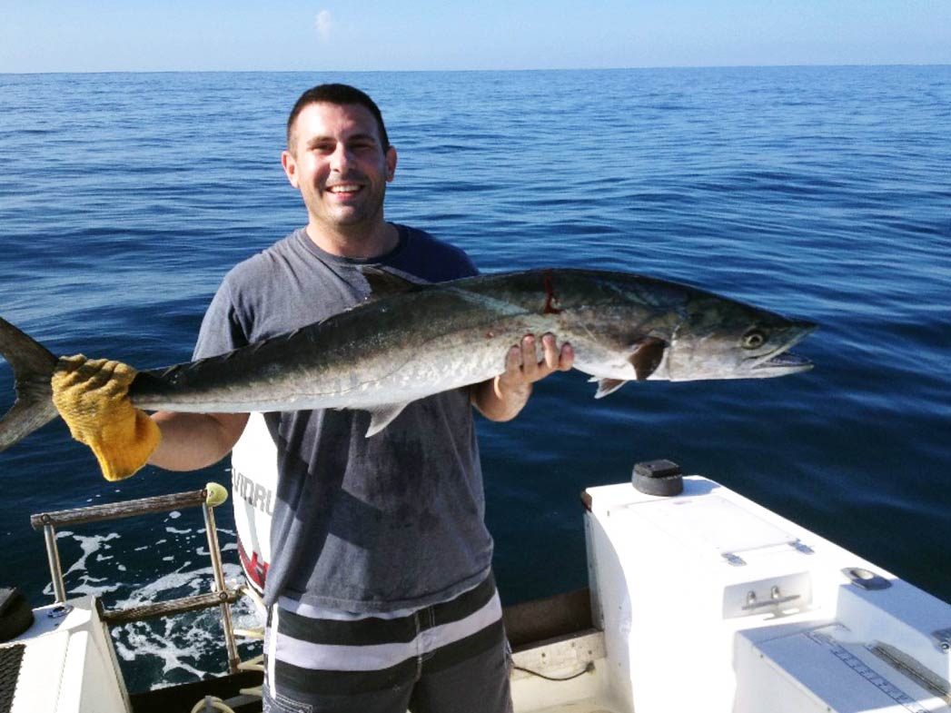 Ein lächelnder Mann steht auf einem Boot und hält eine große Königsmakrele in der Hand, mit den Gewässern des Atlantiks im Rücken