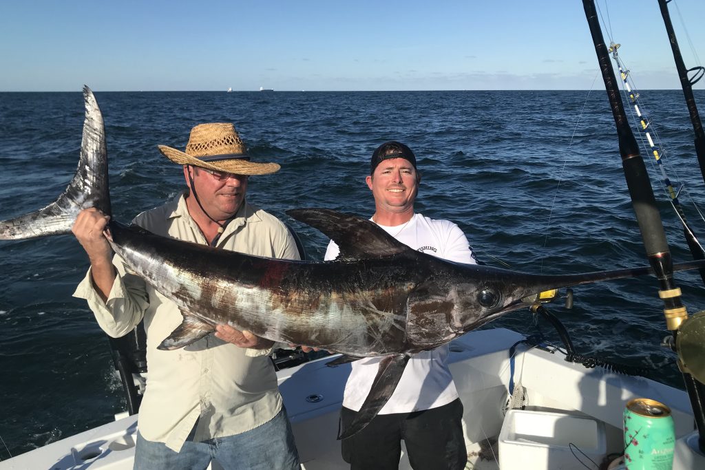 Two fishermen holding a large Swordfish on a charter boat, with water and blue skies in the background