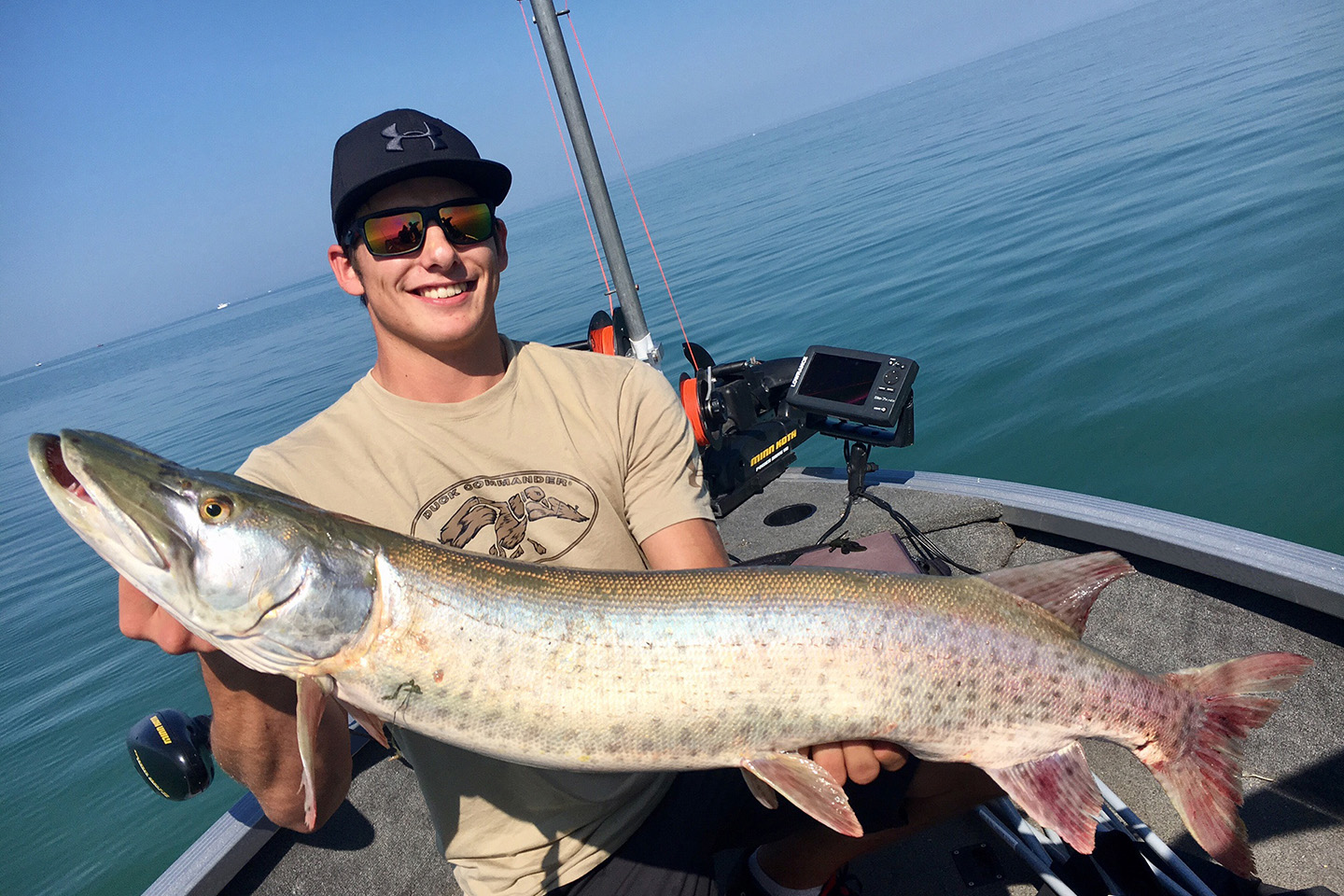 A smiling angler on a boat holding a Muskellunge fish with water behind him.