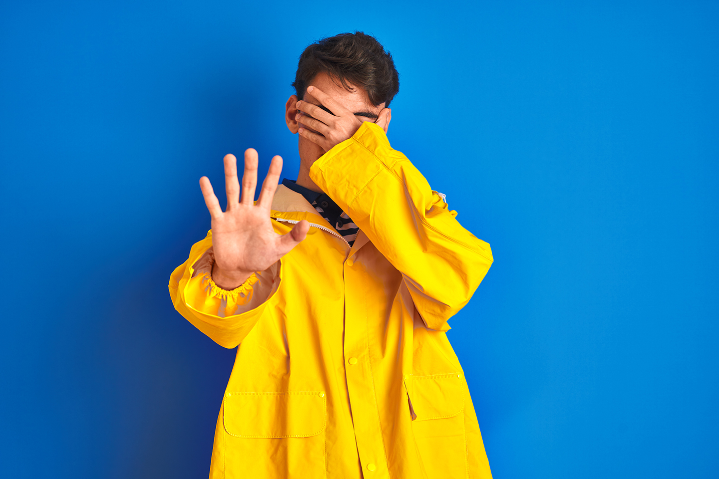 A young man in a yellow fisherman's jacket holding one hand to his face and the other out to the camera