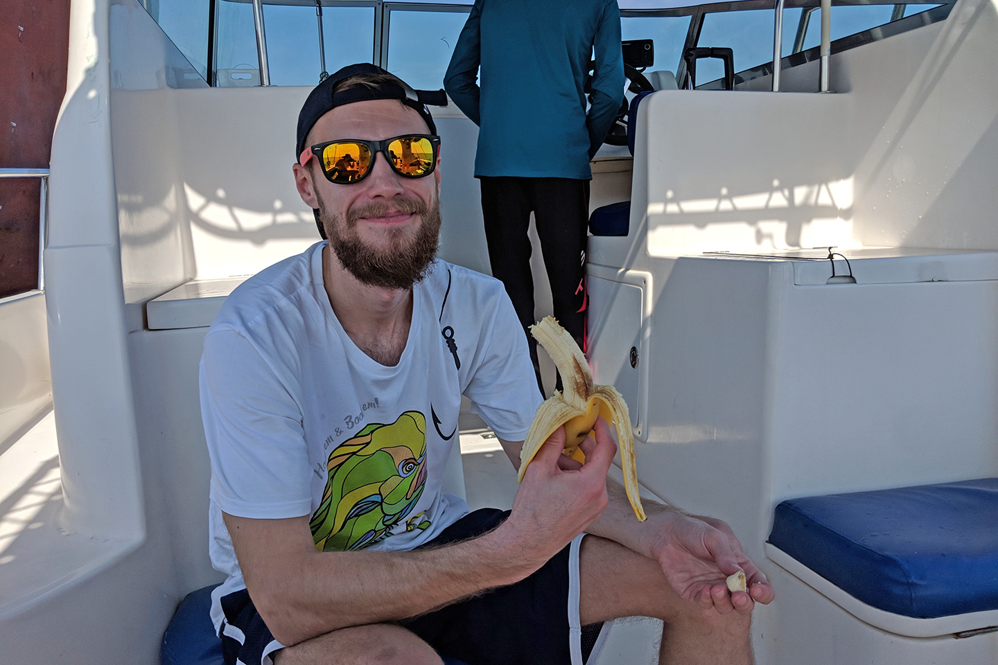 A male angler in a cap and sunglasses eating a banana on a charter fishing boat