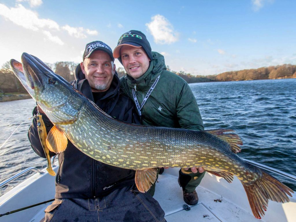 Two anglers wearing winter gear and baseball caps holding a big Northern Pike on a boat, with the water and shoreline behind them