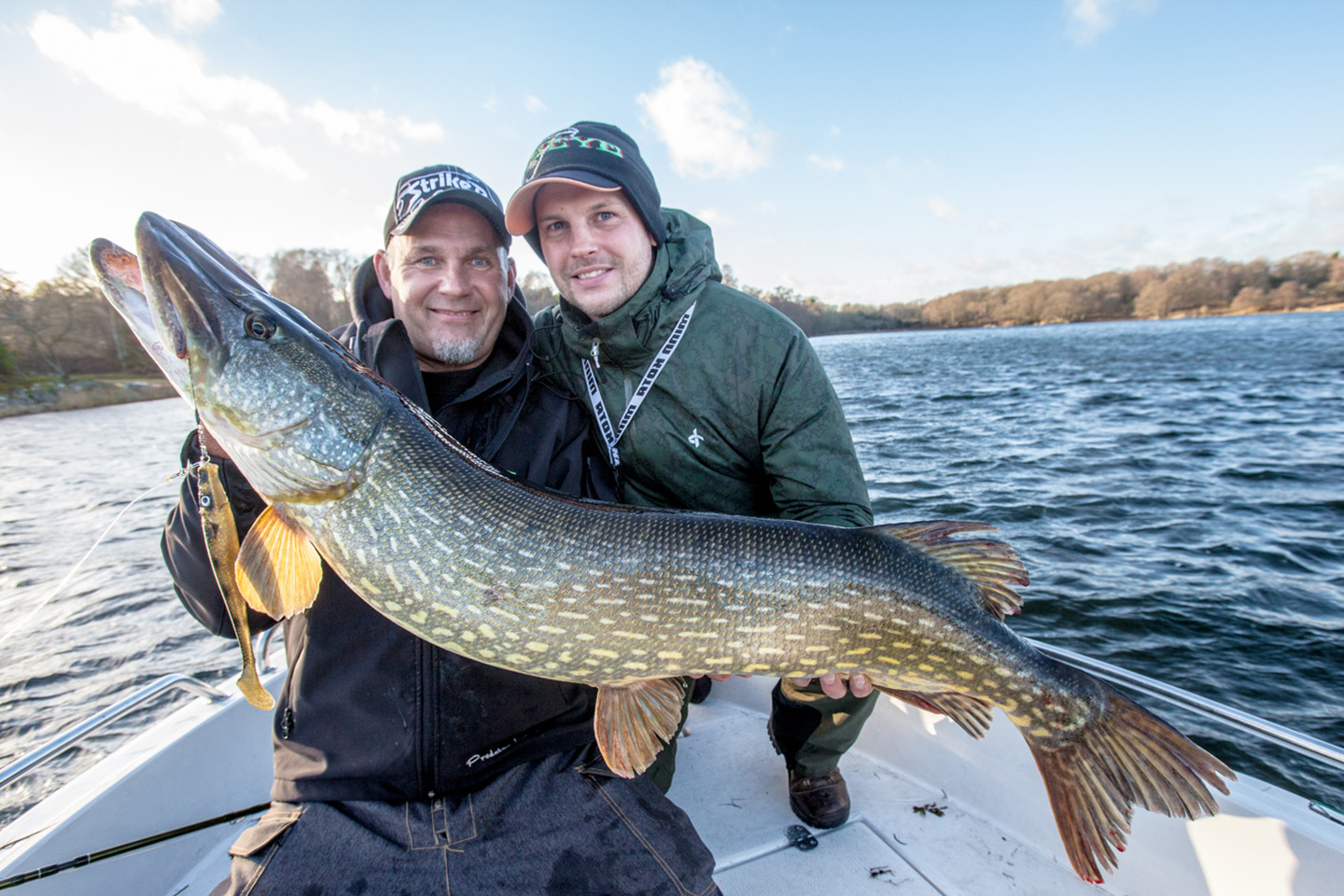 Two anglers on a boat holding a trophy-sized Northern Pike with blue sky and water behind them. The Pike has a large lure handing out of its mouth.