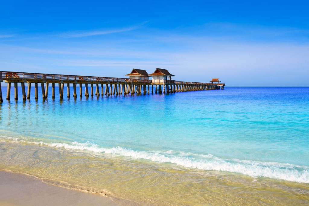 Widok na Naples Pier z plaży, z jasnoniebieską wodą i żółtym piaskiem na pierwszym planie