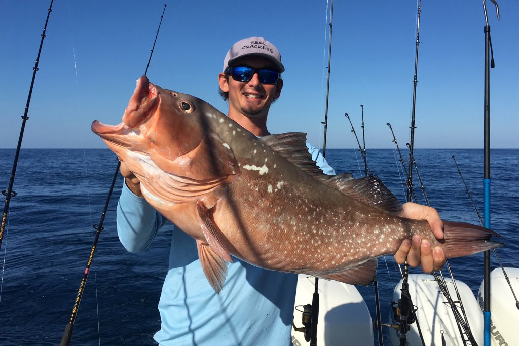 Um pescador sorridente num barco de pesca com um grande Garoupa Vermelha. Há muitas varas de pesca apoiadas atrás dele